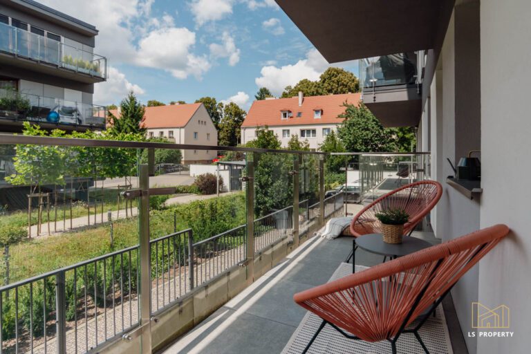 Balkon z dwoma krzesłami i niedużym, okrągłym stolikiem. Widok na okoliczne bloki.