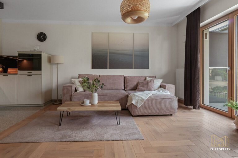 Pokój dzienny z aneksem kuchennym i częścią jadalnianą. Na środku przy ścianie różowa kanapa ze stolikiem kawowym. Z pokoju wyjście na balkon przez duże drzwi tarasowe z oknem.