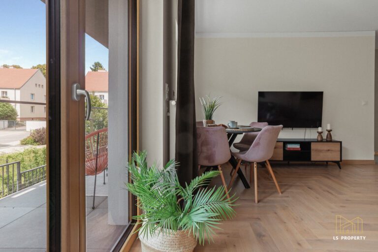 Wyjście na balkon z pokoju dziennego. W rogu pokoju okrągły stolik jadalniany z krzesłami. Na ścianie zawieszony telewizor, pod nim niska szafka.