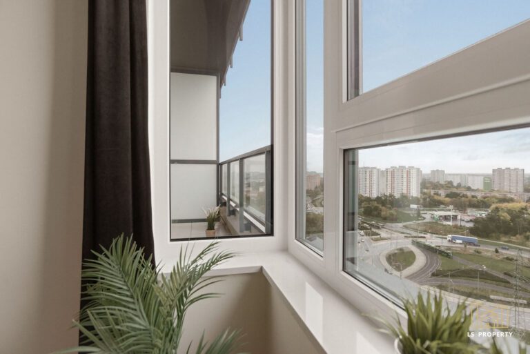 Narożne okno w sypialni wychodzące jednym bokiem na balkon. Z okna widok na panoramę miasta.