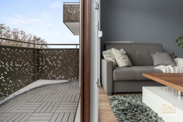 Wyjście na balkon z pokoju dziennego, w którym stoi mała, szara kanapa ze stolikiem kawowym.