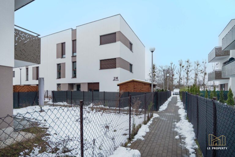 Budynek mieszkalny na osiedlu niskich budynków wielorodzinnych w nowoczesnym stylu. Jasna elewacja z drewnianymi wstawkami. Teren wokół zadbany i ogrodzony.